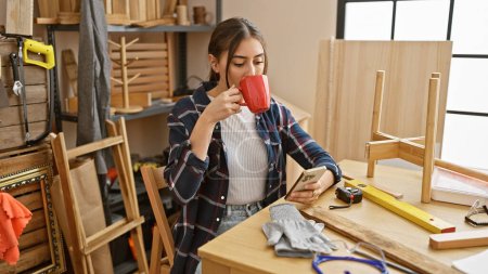 Téléchargez les photos : Femme hispanique aime le café tout en envoyant des textos dans un atelier de travail du bois. - en image libre de droit