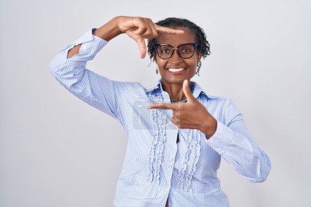 Foto de Mujer africana con rastas de pie sobre fondo blanco con gafas sonrientes haciendo montura con manos y dedos con la cara feliz. concepto de creatividad y fotografía. - Imagen libre de derechos