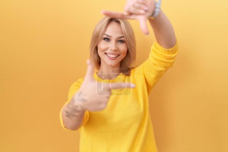 Téléchargez les photos : Jeune femme caucasienne portant un pull jaune souriant faisant cadre avec les mains et les doigts avec le visage heureux. concept de créativité et photographie. - en image libre de droit