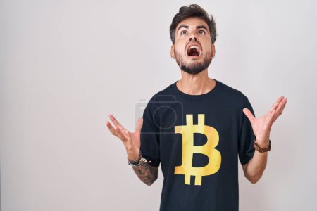 Foto de Joven hombre hispano con tatuajes usando camiseta bitcoin loco y loco gritando y gritando con expresión agresiva y los brazos levantados. concepto de frustración. - Imagen libre de derechos