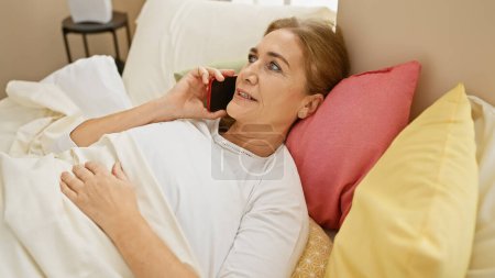 Téléchargez les photos : Une femme caucasienne mature se détend au lit avec un téléphone à la maison, évoquant un sentiment de confort et de connexion. - en image libre de droit