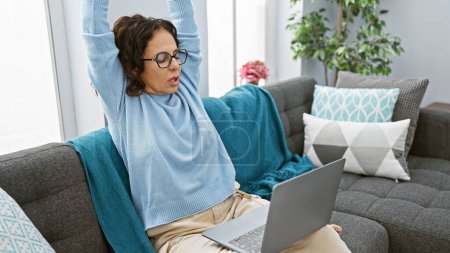 Téléchargez les photos : Une femme mûre s'étirant tout en travaillant sur un ordinateur portable dans un salon confortable. - en image libre de droit