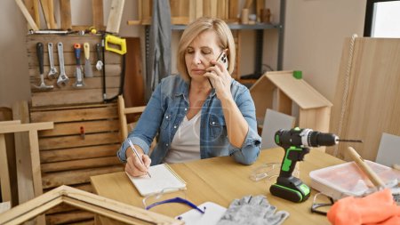 Téléchargez les photos : Une femme d'âge moyen multitâches dans un atelier de menuiserie, parlant au téléphone tout en prenant des notes. - en image libre de droit