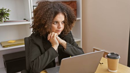 Foto de Una mujer hispana joven y pensativa con el pelo rizado trabajando en una oficina moderna, portátil y café en el escritorio. - Imagen libre de derechos