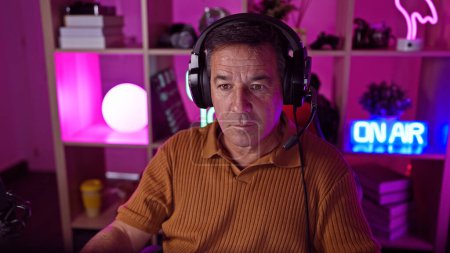 Téléchargez les photos : Homme sérieux d'âge moyen avec écouteurs dans une salle de jeux colorée la nuit transmet une atmosphère de loisirs vibrante. - en image libre de droit