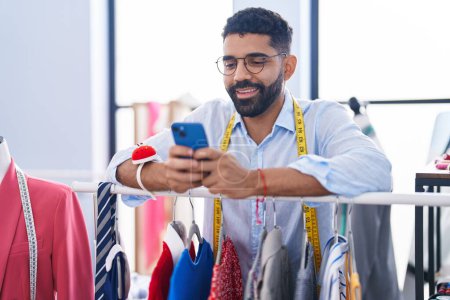 Téléchargez les photos : Jeune tailleur arabe utilisant un smartphone appuyé sur un porte-vêtements au magasin de tailleurs - en image libre de droit