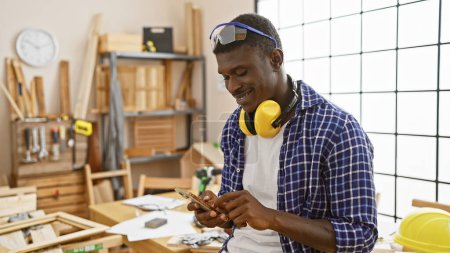 Photo for An african american man in a plaid shirt smiles while using a smartphone in a well-equipped carpentry workshop. - Royalty Free Image