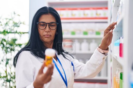 Téléchargez les photos : Middle age hispanic woman pharmacist holding pills bottle on shelving at pharmacy - en image libre de droit