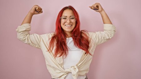 Photo for Joyful young redhead sportswoman confidently flexing arms, flaunting power & happiness over isolated pink background, a portrait of cheerful, sporty confidence. - Royalty Free Image