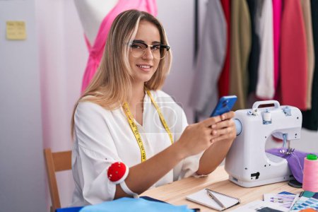Téléchargez les photos : Jeune femme blonde tailleur souriant confiant en utilisant smartphone au magasin de vêtements - en image libre de droit