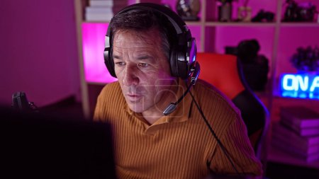 Photo for Mature man with headphones in gaming room illuminated by neon lights focused on computer screen - Royalty Free Image