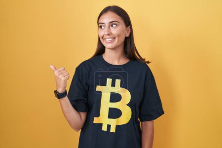 Téléchargez les photos : Jeune femme hispanique portant Bitcoin t-shirt pointant vers le dos derrière avec la main et les pouces vers le haut, souriant confiant - en image libre de droit