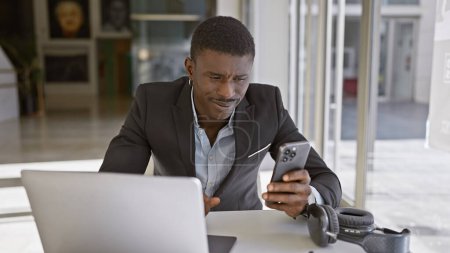 Photo for A professional african man in a suit working with a laptop and smartphone in a modern office setting - Royalty Free Image