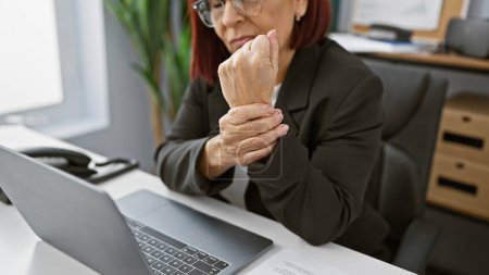 Mature hispanic woman with glasses in pain at office, holding wrist in discomfort by laptop and paperwork.