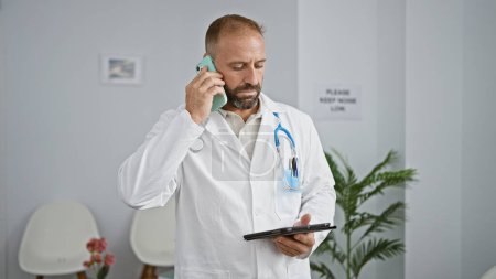 Photo for Young man doctor using touchpad talking on smartphone at clinic waiting room - Royalty Free Image