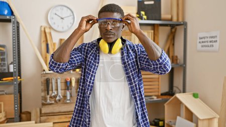 Photo for African american man wearing safety goggles in a carpentry workshop setting up for work. - Royalty Free Image
