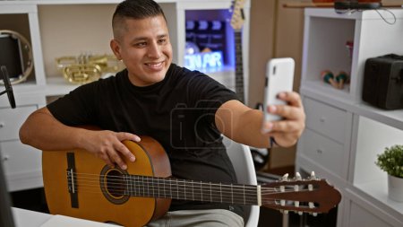 Photo for Smiling young latin man, an artist in his prime, striking a melody on his classical guitar, takes a selfie with his smartphone in a music studio - Royalty Free Image