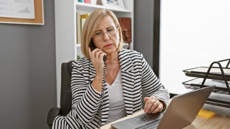 Foto de Una mujer madura en una oficina hablando por teléfono y trabajando en un ordenador portátil, retratando un entorno profesional ocupado. - Imagen libre de derechos