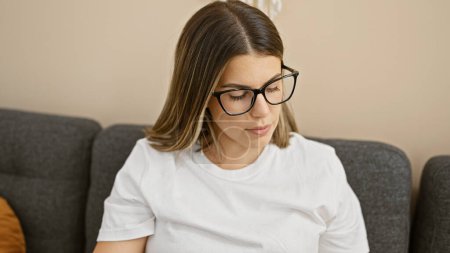 Photo for Hispanic woman in glasses sitting thoughtfully in a neutral-toned living room. - Royalty Free Image