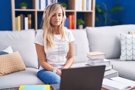 Téléchargez les photos : Jeune femme blonde étudiant à l'aide d'un ordinateur portable à la maison détendue avec une expression sérieuse sur le visage. simple et naturel regardant la caméra. - en image libre de droit