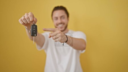 Foto de Joven hombre hispano sonriendo confiado señalando la llave de un coche nuevo sobre un fondo amarillo aislado - Imagen libre de derechos