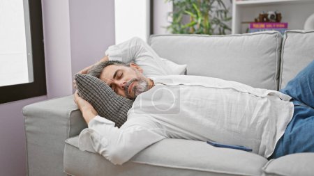 Jeune homme hispanique épuisé avec une belle barbe aux cheveux gris confortablement couché sur le canapé confortable, dormant profondément et profitant de la détente dans le salon de sa maison, échappant à la réalité fatiguée.