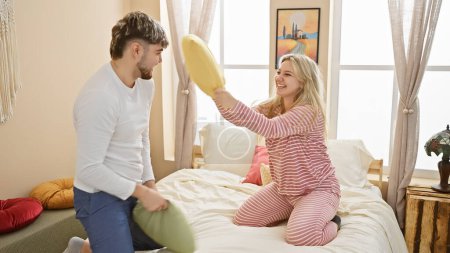 A playful couple engages in a pillow fight in a cozy bedroom, capturing love and joy in a domestic setting.