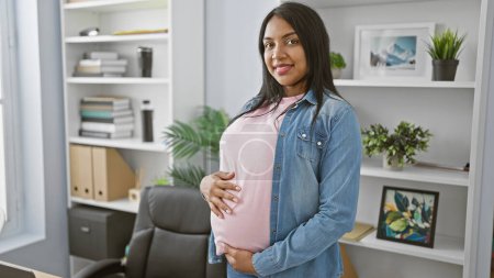 Photo for Smiling young pregnant woman confidently touches belly while working in the office, embodying maternity and business success. - Royalty Free Image