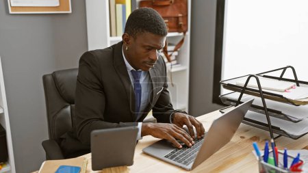 Foto de Un hombre de negocios afroamericano se centra intensamente en su computadora portátil en un entorno de oficina moderno. - Imagen libre de derechos