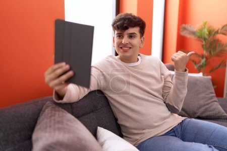 Téléchargez les photos : Jeune homme non binaire utilisant le pavé tactile assis sur le canapé pointant le pouce vers le côté souriant heureux avec la bouche ouverte - en image libre de droit