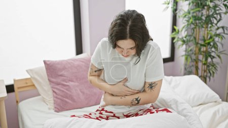 Photo for Young woman with tattoos experiencing stomach pain while sitting on a bed in her bedroom. - Royalty Free Image