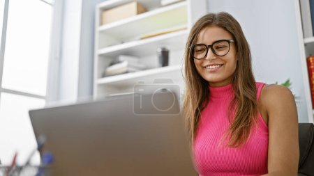 Photo for Confident young hispanic woman, donned in an elegant attire, beaming with joy as she works on her laptop at her office desk, exuding professional success - Royalty Free Image