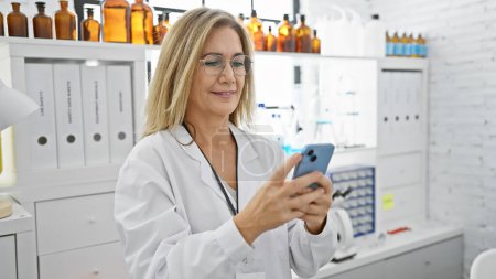 Photo for A mature blonde woman in a lab coat uses a smartphone in a modern laboratory setting, surrounded by equipment and glassware. - Royalty Free Image
