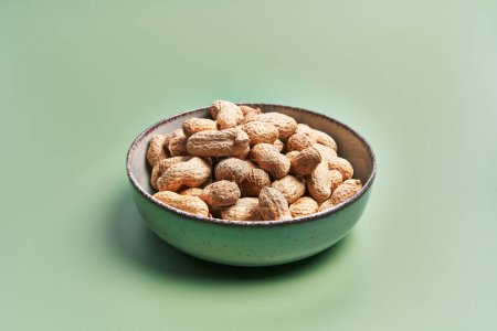 Foto de Un tazón lleno de cacahuetes sin cáscara presentado sobre un fondo verde liso, que representa la simplicidad y los conceptos de alimentos orgánicos. - Imagen libre de derechos