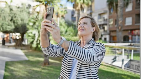 Téléchargez les photos : Femme blonde mature profitant d'un selfie dans un parc ensoleillé - en image libre de droit