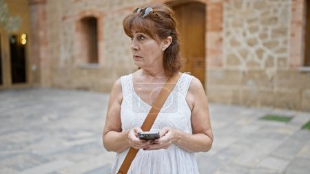Photo for A middle-aged redhead woman in casual attire uses a smartphone on an urban street, embodying a sense of everyday life technology use. - Royalty Free Image