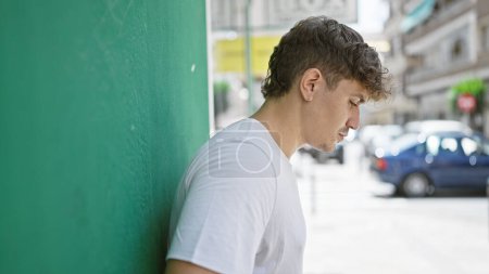 Foto de Joven hombre hispano fresco y relajado apoyado en la pared urbana de la ciudad, su hermoso retrato adulto brillando bajo el sol al aire libre. mirando por la calle de la ciudad, su expresión seria, concentrada. - Imagen libre de derechos