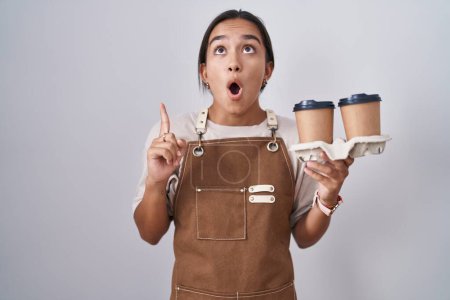 Foto de Mujer hispana joven con delantal de camarera profesional sosteniendo café asombrada y sorprendida mirando hacia arriba y señalando con los dedos y los brazos levantados. - Imagen libre de derechos