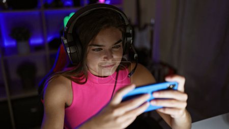 Téléchargez les photos : Jeune joueuse hispanique confiante, portrait d'une belle streamer jouant à un jeu vidéo sur son smartphone, à l'intérieur, dans une salle de jeux sombre - en image libre de droit