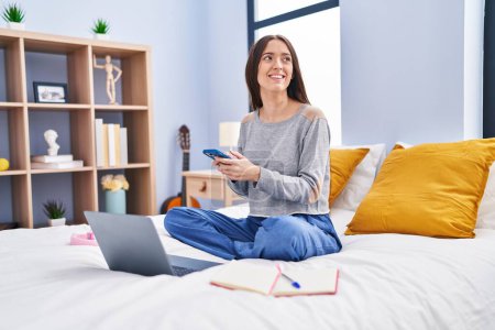 Photo for Young beautiful hispanic woman student using smartphone studying on bed at bedroom - Royalty Free Image