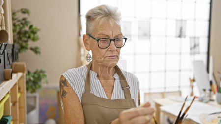 Photo for Dedicated senior grey-haired woman artist, holding paintbrushes, deeply engrossed in painting at art studio. - Royalty Free Image