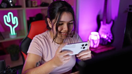 Téléchargez les photos : Femme hispanique souriante utilisant un smartphone dans une salle de jeux colorée éclairée au néon à la maison la nuit - en image libre de droit