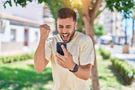 Téléchargez les photos : Jeune homme hispanique utilisant un smartphone avec expression gagnante au parc - en image libre de droit