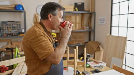 Foto de Hombre maduro bebiendo café en un taller de carpintería bien equipado, que representa el tiempo de descanso de un artesano. - Imagen libre de derechos