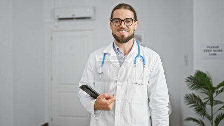 Téléchargez les photos : Jeune homme hispanique médecin tenant pavé tactile souriant à la salle d'attente clinique - en image libre de droit