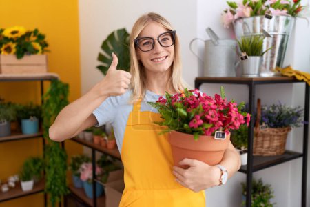 Téléchargez les photos : Jeune femme caucasienne travaillant au magasin de fleuriste tenant l'usine souriant heureux et positif, pouce vers le haut faisant l'excellent et signe d'approbation - en image libre de droit