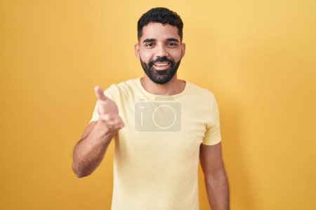 Téléchargez les photos : Homme hispanique avec barbe debout sur fond jaune souriant joyeuse offrant la main de la paume donnant assistance et acceptation. - en image libre de droit