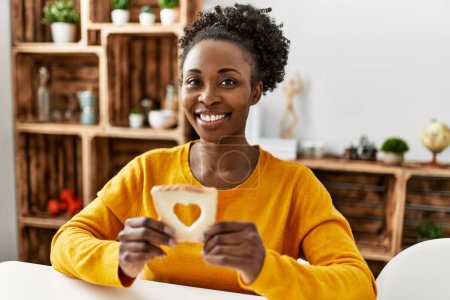 Photo for African american woman holding bread with heart hole shape sitting on table at home - Royalty Free Image