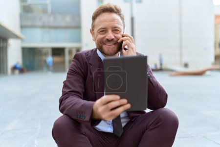Photo for Middle age man business worker using touchpad talking on smartphone at street - Royalty Free Image