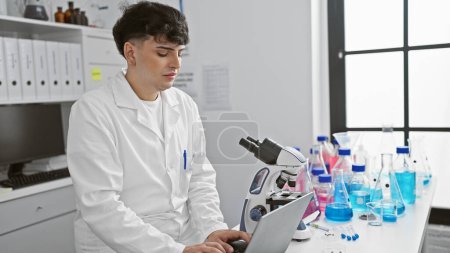 Photo for A young man in a white lab coat works intently on a laptop in a bright laboratory, with a microscope and scientific equipment around. - Royalty Free Image